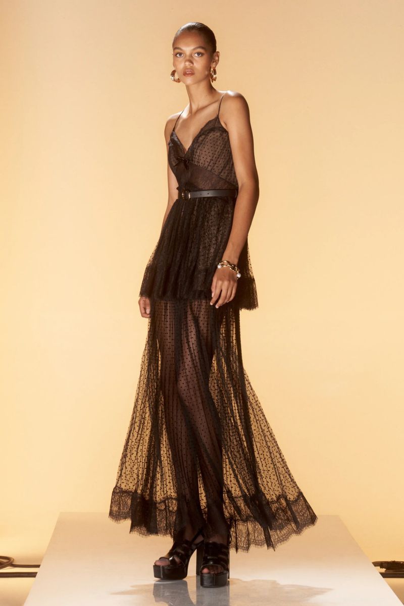 Full-length shot of a young woman in a black dress with spaghetti straps, a thin belt, and platform shoes on a white platform.