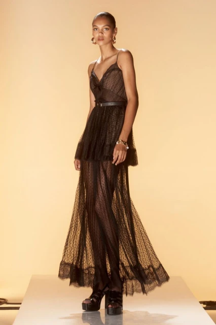 Full-length shot of a young woman in a black dress with spaghetti straps, a thin belt, and platform shoes on a white platform.