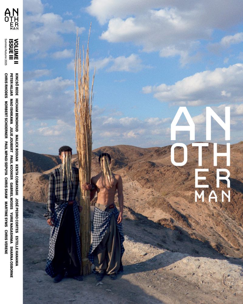Magazine cover featuring two men in stylized desert fashion, holding tall straw bundle against a backdrop of desert hills and blue sky.