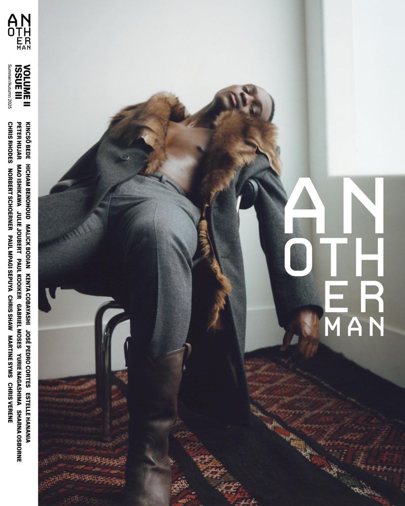 Black male model in a fur-trimmed grey overcoat and leather boots reclining on a chair for AnOther Man Magazine cover.