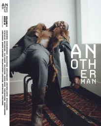 Black male model in a fur-trimmed grey overcoat and leather boots reclining on a chair for AnOther Man Magazine cover.