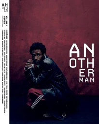 Magazine cover of a Black man crouching in a black leather coat and red track pants against a textured burgundy backdrop.