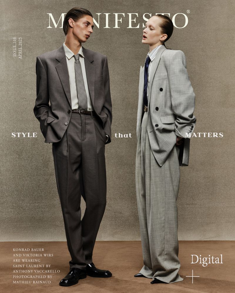 Two fashion models wearing grey suits pose for a magazine cover. The male model wears a suit and tie, while the female model wears a double-breasted suit.