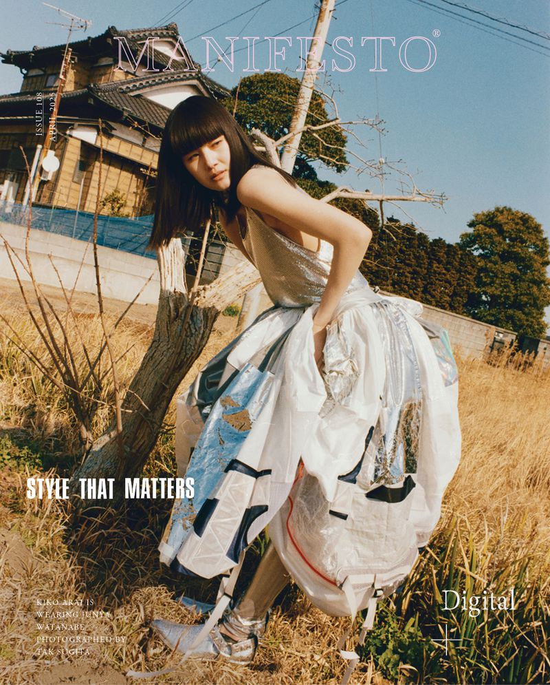 Editorial fashion shot of a Japanese woman in a silver top and sculptural skirt, posing outdoors with a traditional house backdrop.