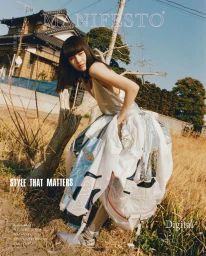 Model wearing a voluminous silver and white dress leaning against a tree in a dry grassy field before a traditional house