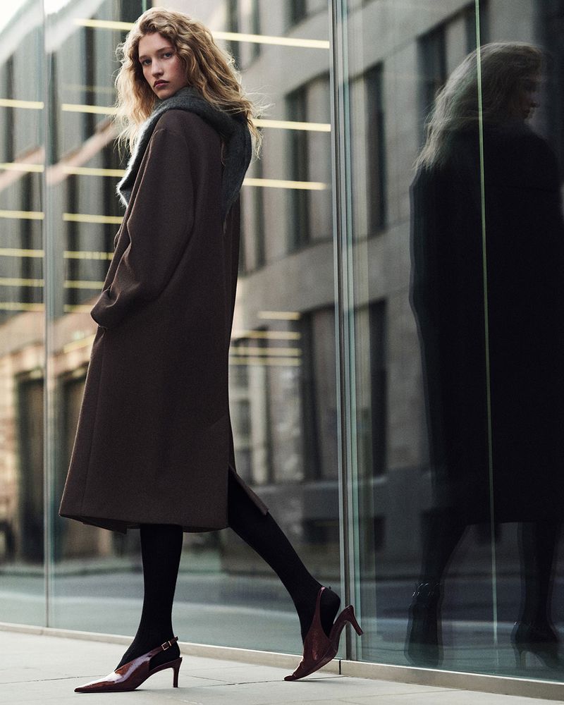 A stylish woman wearing a long brown coat, black tights, and high heels, stands on a city sidewalk with a mirrored building behind.