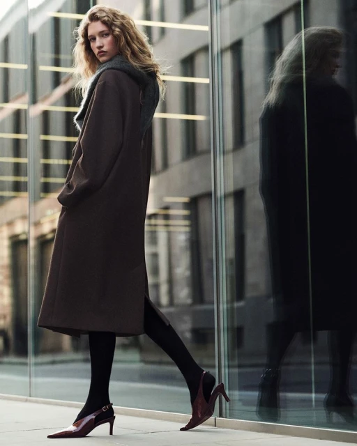 A stylish woman wearing a long brown coat, black tights, and high heels, stands on a city sidewalk with a mirrored building behind.