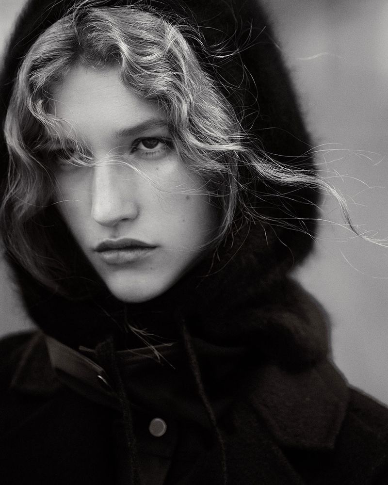 Close-up monochrome portrait of a woman wearing a dark hooded coat with wisps of curly hair blowing across her face.