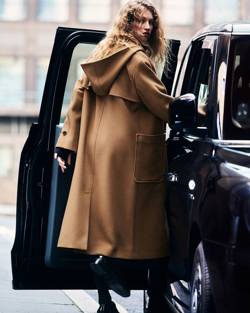 A woman with long, wavy hair and a tan hooded duffle coat exits a black car in an urban environment. The coat is a focal point.
