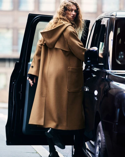 A woman with long, wavy hair and a tan hooded duffle coat exits a black car in an urban environment. The coat is a focal point.