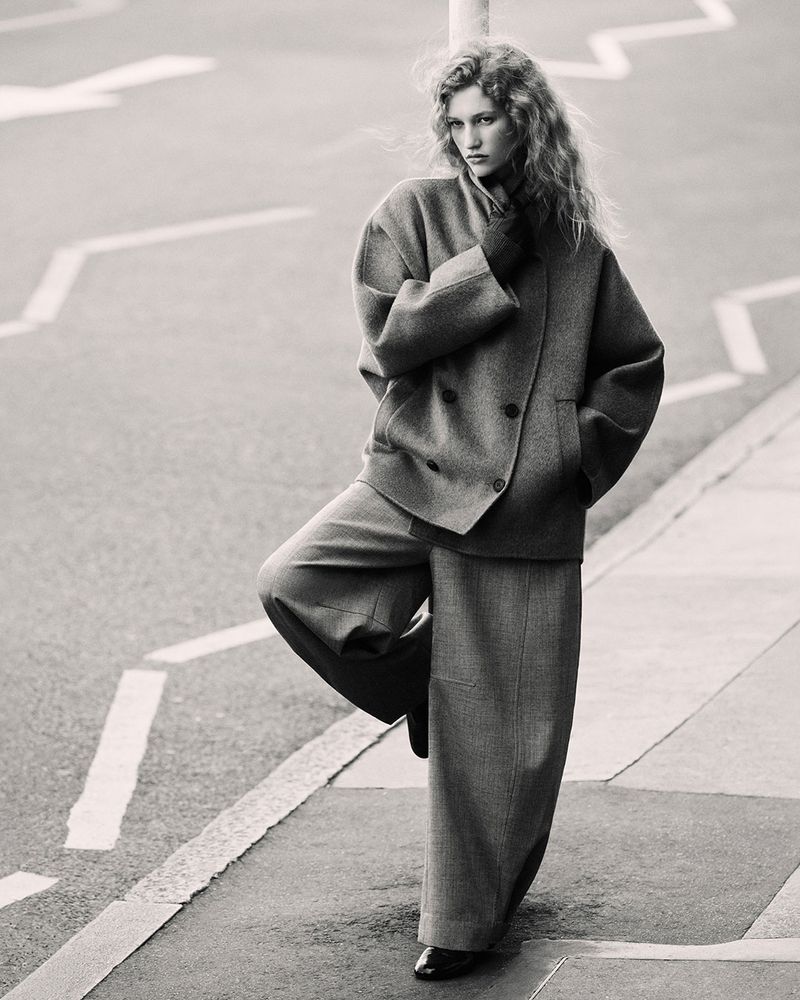 Adult female model in double-breasted wool coat and wide-leg pants poses on street corner against gray monochrome background in studio shot.