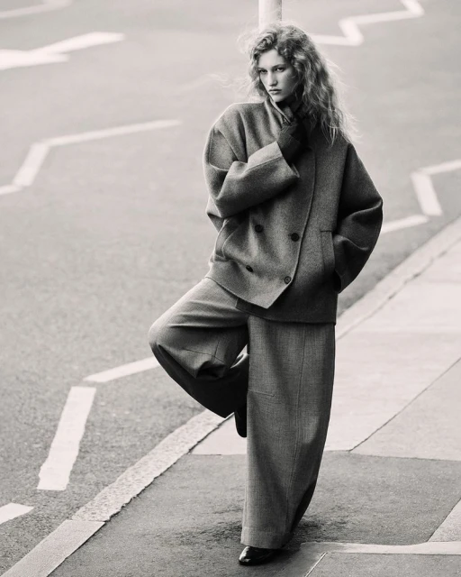 Adult female model in double-breasted wool coat and wide-leg pants poses on street corner against gray monochrome background in studio shot.