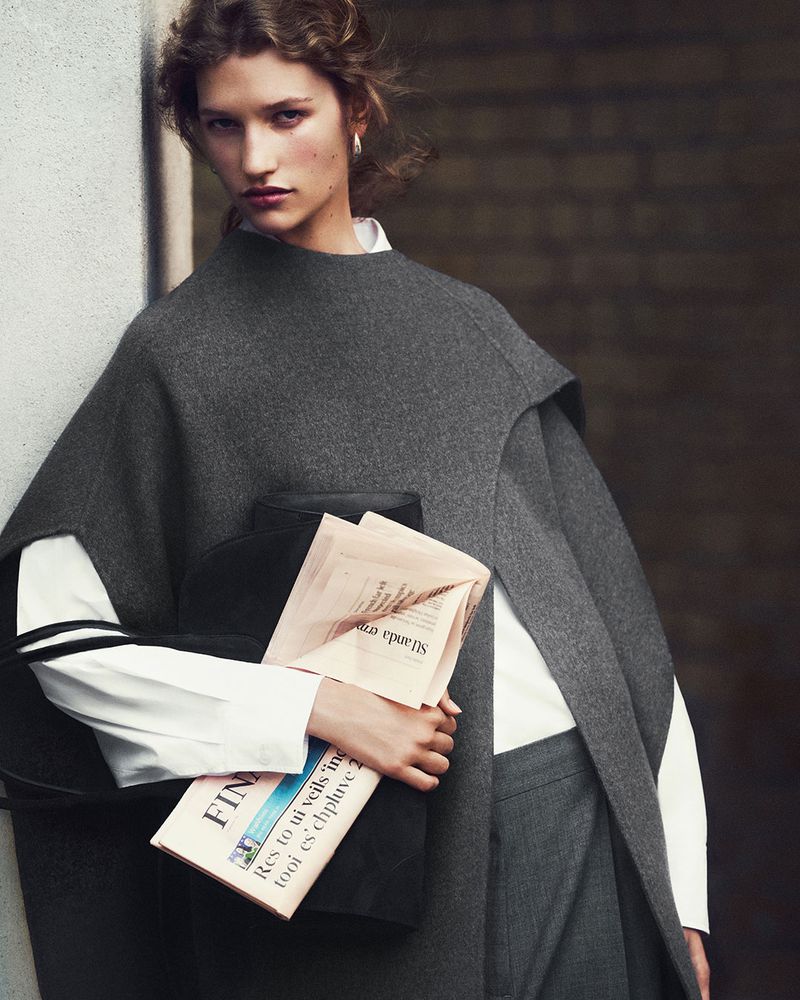 A woman in a gray wool outfit holds a newspaper and black clutch, posing against a muted background in a fashion editorial shot.