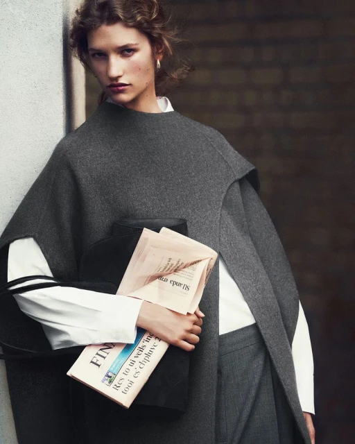 A woman in a gray wool outfit holds a newspaper and black clutch, posing against a muted background in a fashion editorial shot.
