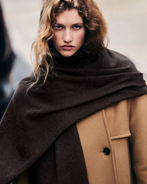 Portrait of an adult woman wearing a brown scarf and a two-tone tan and brown coat, posing outdoors with a neutral expression.