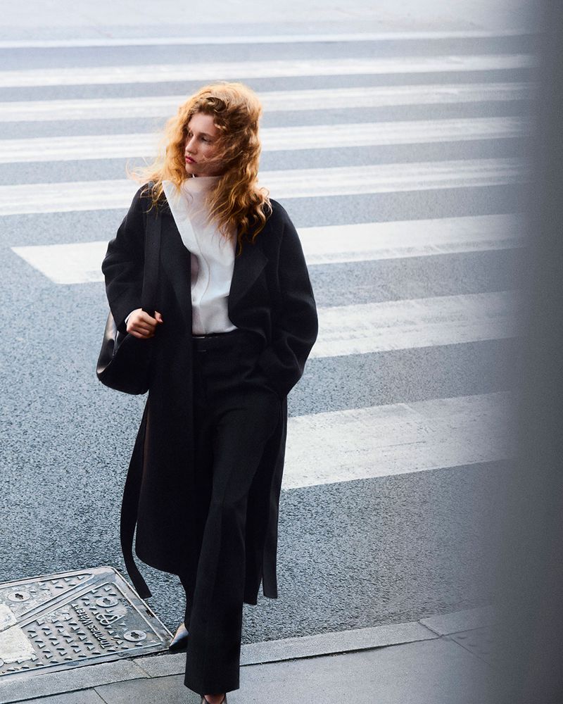 A redhead woman in a black coat and trousers walks across a zebra crossing on a city street, carrying a shoulder bag in daylight.