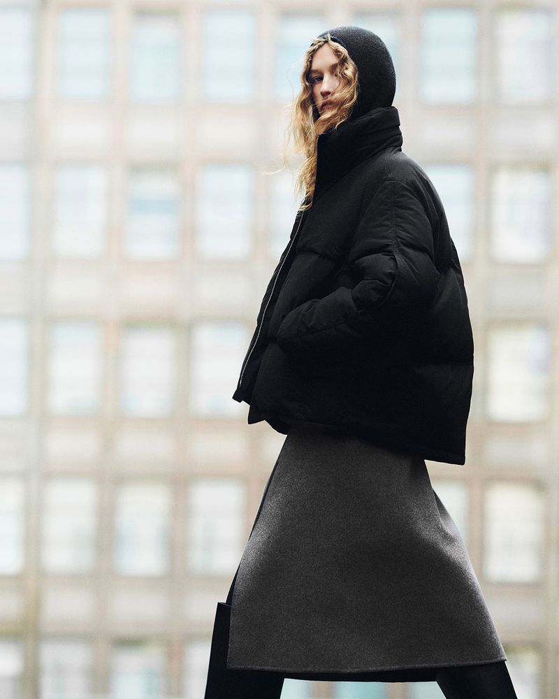An adult woman wearing a black puffer jacket with a hood and a grey skirt is walking outdoors with a blurred background of city buildings.