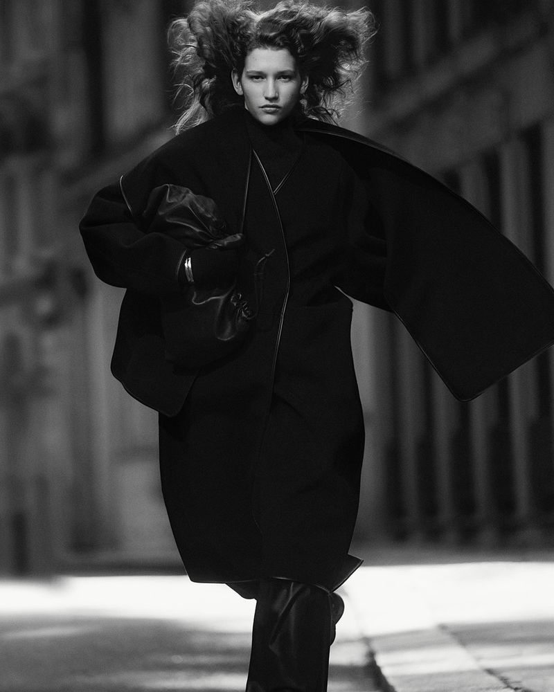Fashionable woman in motion wearing a black coat, gloves, and carrying a black clutch on a city street, captured in black and white.