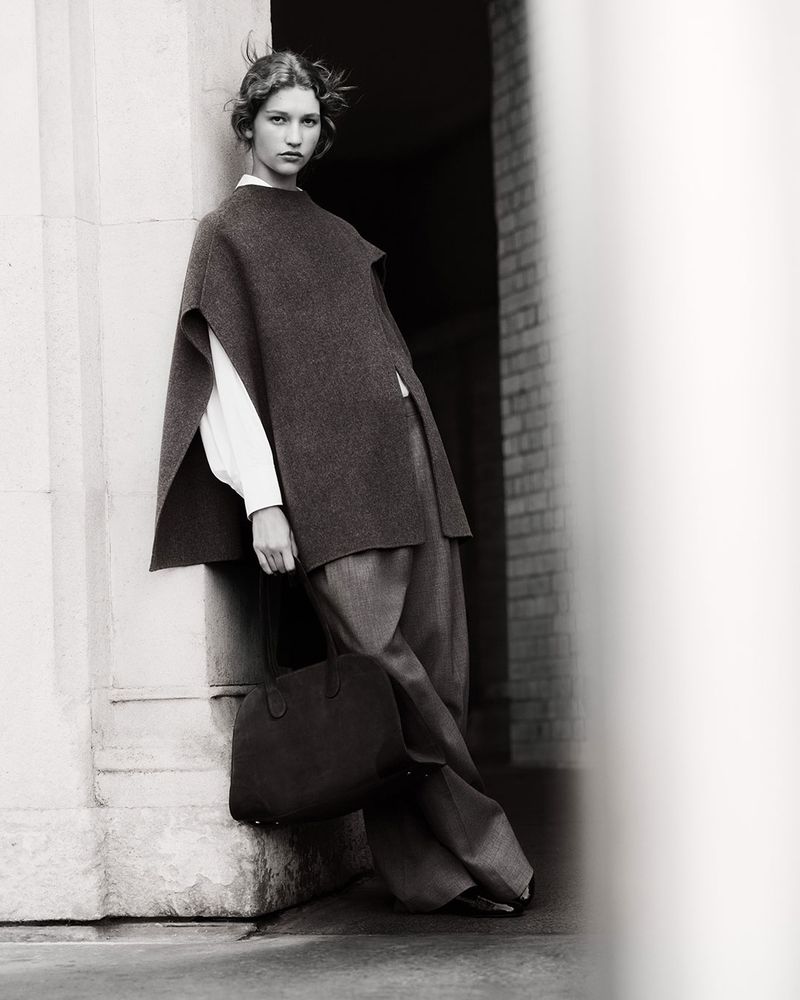 Monochrome fashion shot of a woman in a wool poncho, trousers, and a tote bag, leaning against a column in a building entrance.