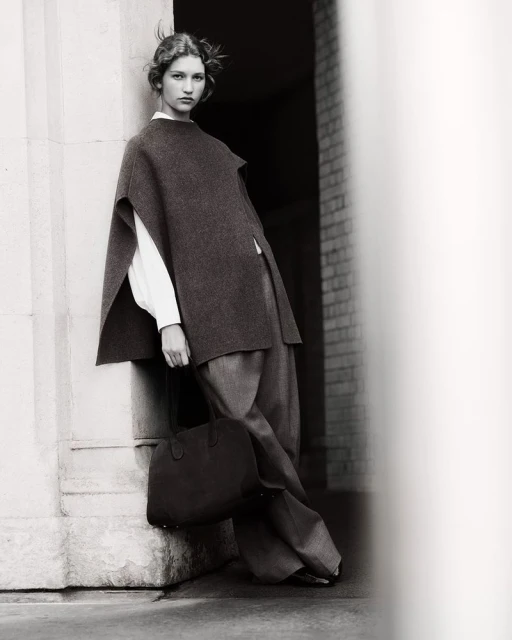 Monochrome fashion shot of a woman in a wool poncho, trousers, and a tote bag, leaning against a column in a building entrance.