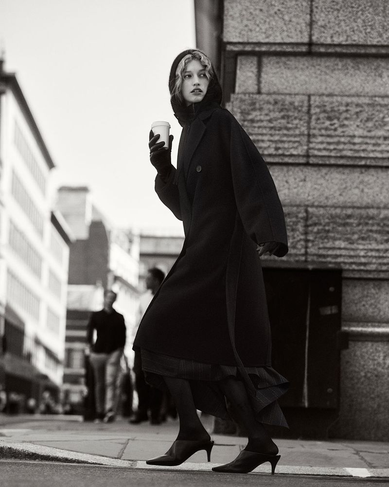 Monochrome fashion shot of a stylish young woman in a black coat walking on a city street with a coffee cup on a cloudy day.
