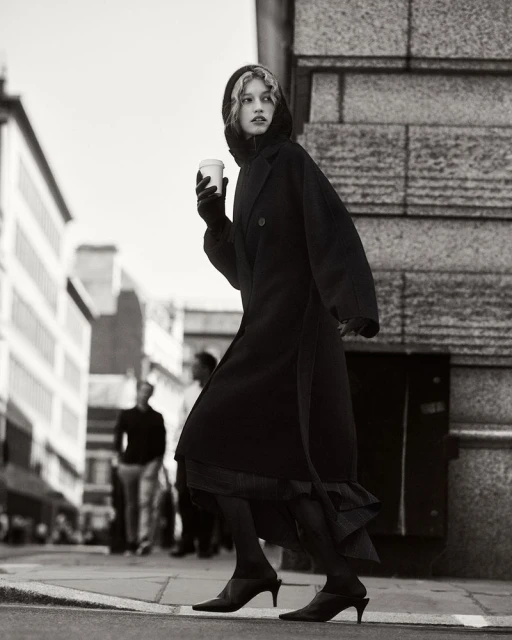 Monochrome fashion shot of a stylish young woman in a black coat walking on a city street with a coffee cup on a cloudy day.