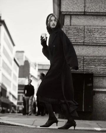Monochrome fashion shot of a stylish young woman in a black coat walking on a city street with a coffee cup on a cloudy day.