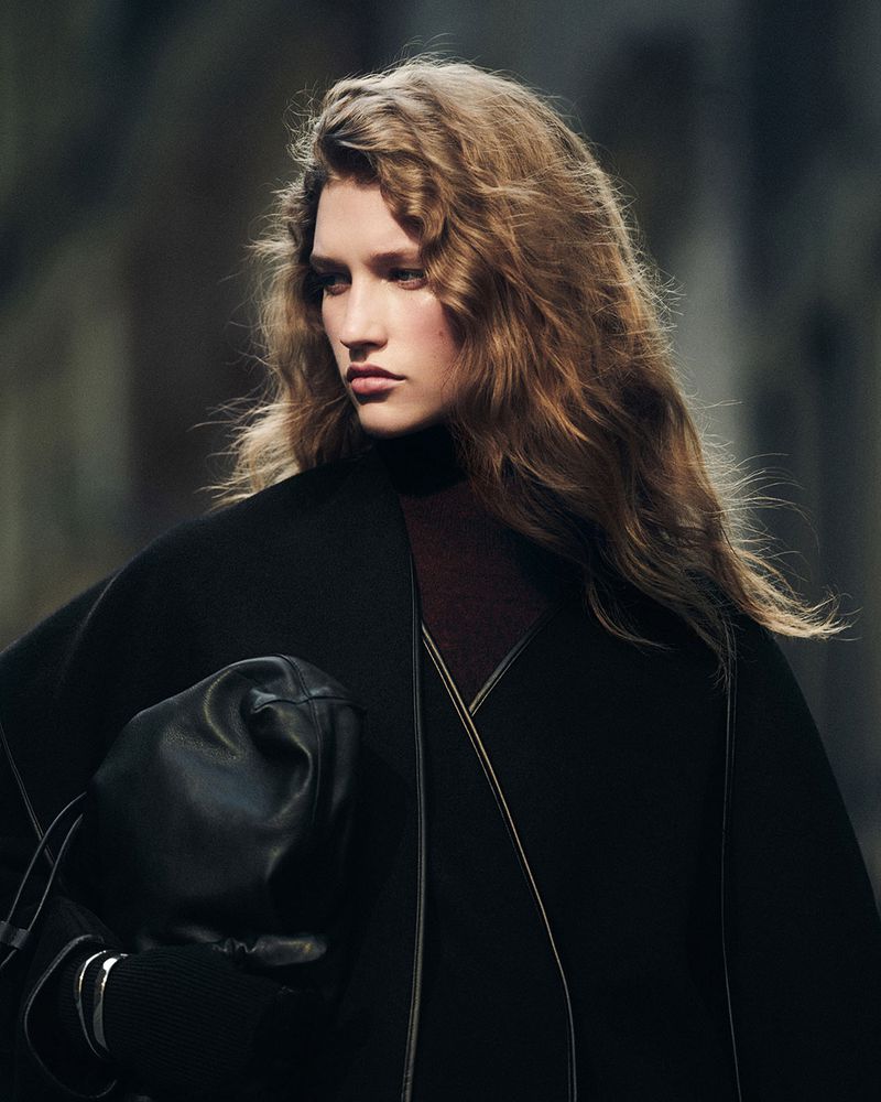 A fair-skinned woman with wavy brown hair wears a dark cape and carries a black leather bag in a fashion shot, set against an urban backdrop.