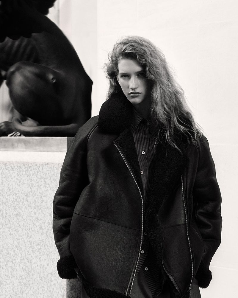 A woman in a dark jacket with a fur collar poses next to a stone statue; the image is black and white, emphasizing textures and shadows.