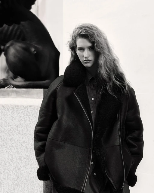 A woman in a dark jacket with a fur collar poses next to a stone statue; the image is black and white, emphasizing textures and shadows.