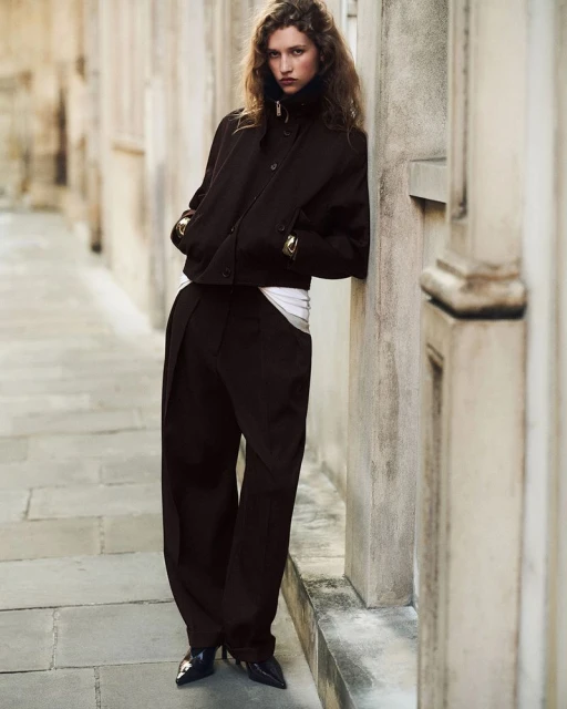 Fashionable adult woman in dark jacket and wide-leg trousers leaning against a light stone wall in an outdoor urban setting.