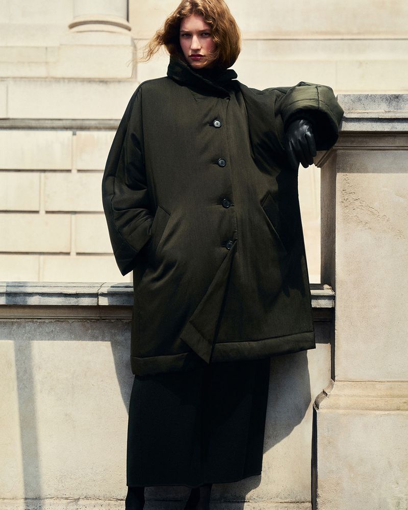 Model wearing a dark green padded coat leaning against a stone building exterior