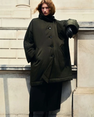 Model wearing a dark green padded coat leaning against a stone building exterior