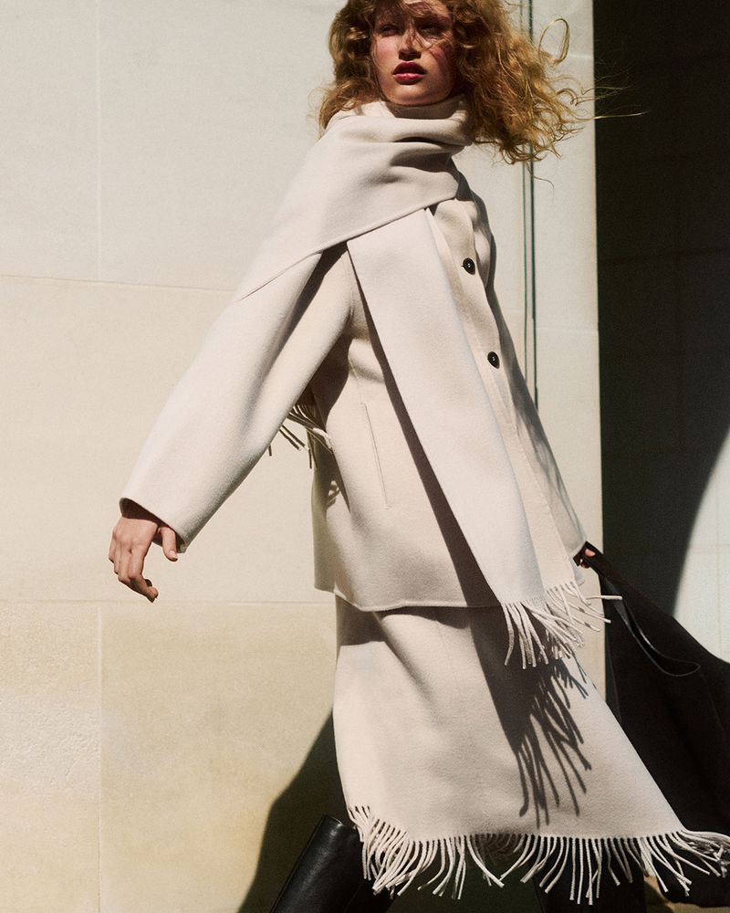 Model with curly blonde hair wearing a cream-colored fringed wool scarf and coat against a stone wall.