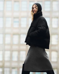 Model in a black puffer jacket with a hood and a grey skirt standing against an urban building exterior backdrop.