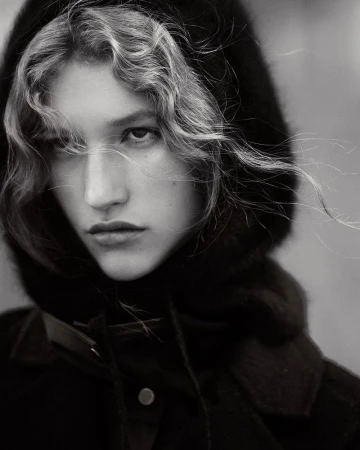 Close-up monochrome portrait of a woman wearing a dark hooded coat with wisps of curly hair blowing across her face.