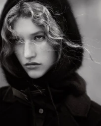 Close-up monochrome portrait of a woman wearing a dark hooded coat with wisps of curly hair blowing across her face.
