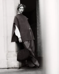 Model leaning against a stone pillar in a dark poncho and trousers holding a handbag for COS Outerwear 2024