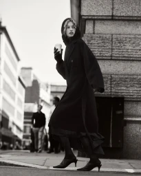 Model wearing a black coat and hooded scarf holding a coffee cup on a city street, photographed by Bryan Liston for COS.