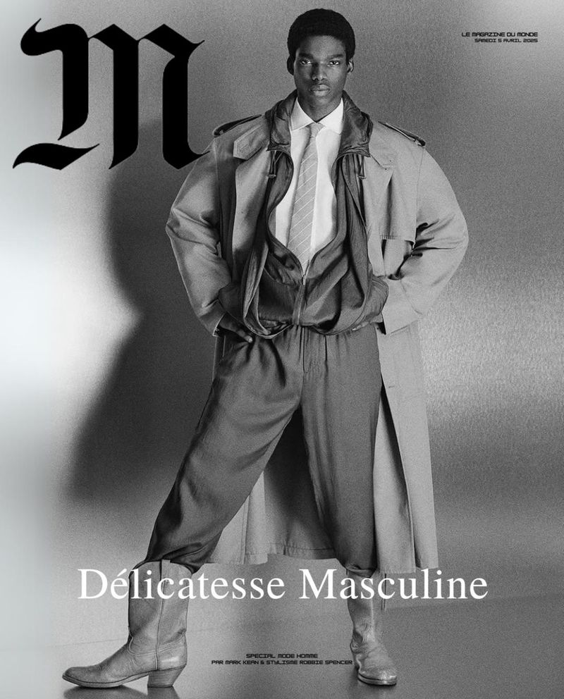 Black male fashion model in trench coat, shirt, tie, and boots poses for a black and white studio shot, showcasing masculine elegance.