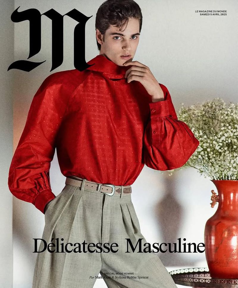Magazine cover featuring a young man in a red blouse and grey trousers, accessorized with a belt, posing against a neutral backdrop with flowers.