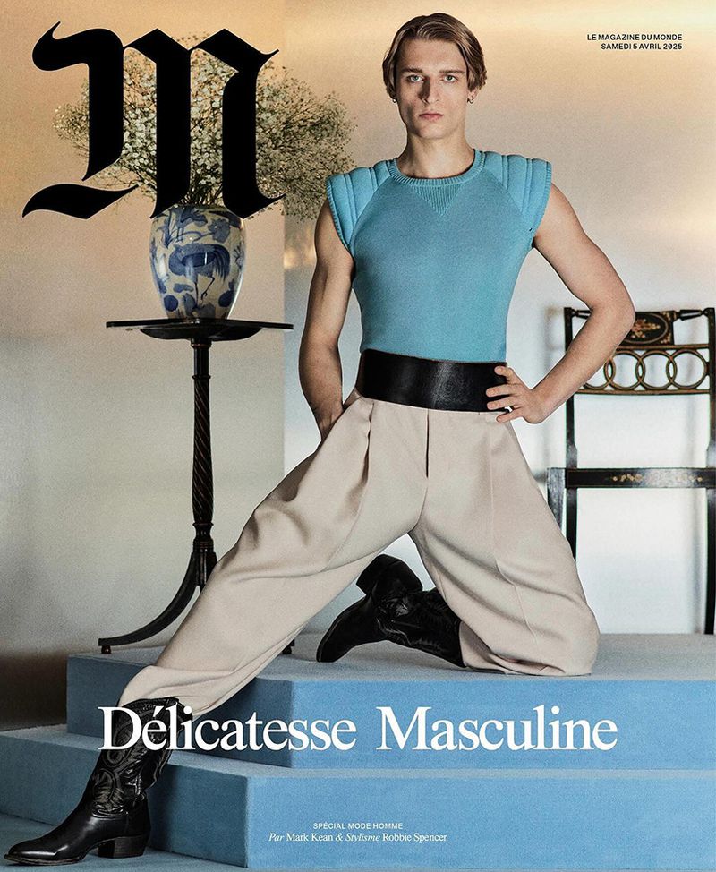 Magazine cover featuring a male model in a blue tank top, wide beige pants, and black cowboy boots, posing on a blue staircase.