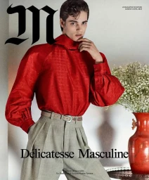 Male model wearing a bright red high-neck puff-sleeve blouse and pleated grey trousers next to a red vase of white flowers.