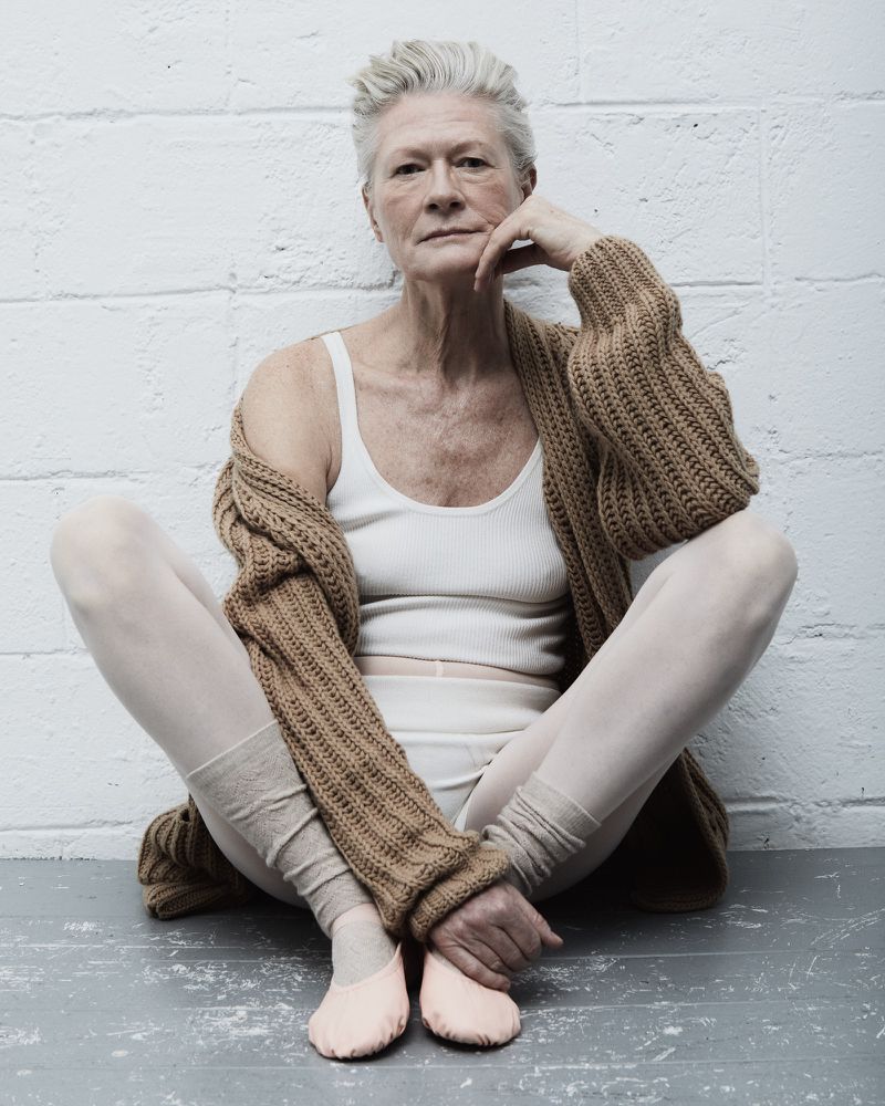 Elderly woman with short grey hair, wearing a cardigan and ballet shoes, sitting in a ballet pose in front of a white brick wall.