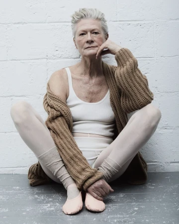 Elderly woman with short grey hair, wearing a cardigan and ballet shoes, sitting in a ballet pose in front of a white brick wall.