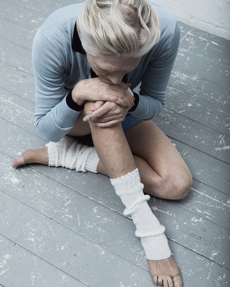 Elderly woman with fair skin sits on gray wood floor in pale blue sweater and white knit leg warmers with her head bent toward her knees.