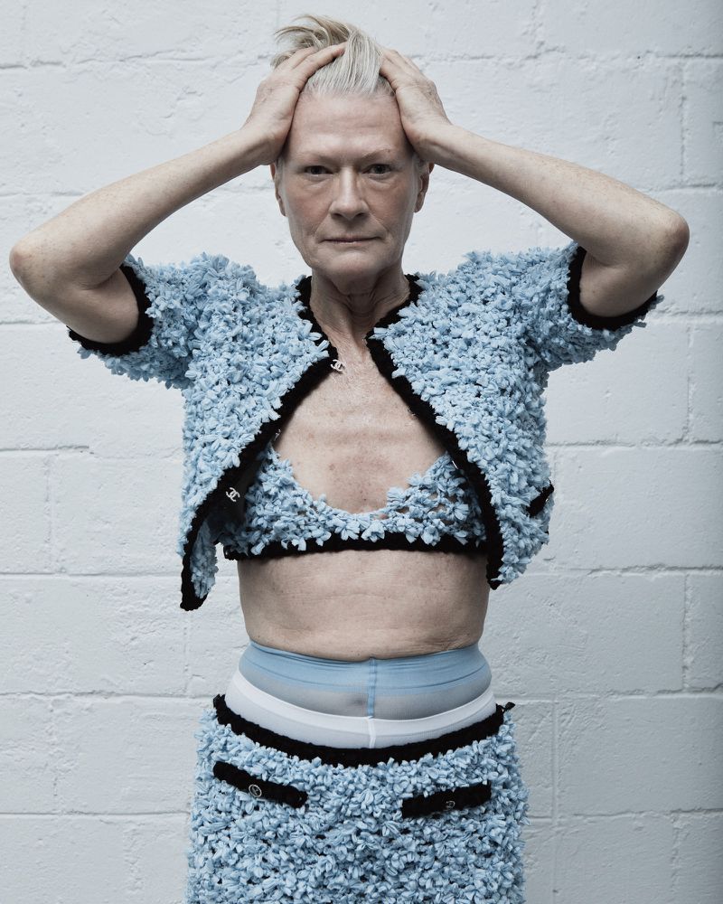 Elderly woman in blue knitwear suit poses in front of white brick wall, hands atop her head, looking forward in a studio shot.