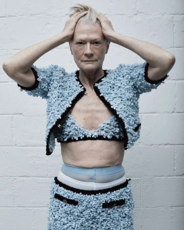 Elderly woman in blue knitwear suit poses in front of white brick wall, hands atop her head, looking forward in a studio shot.