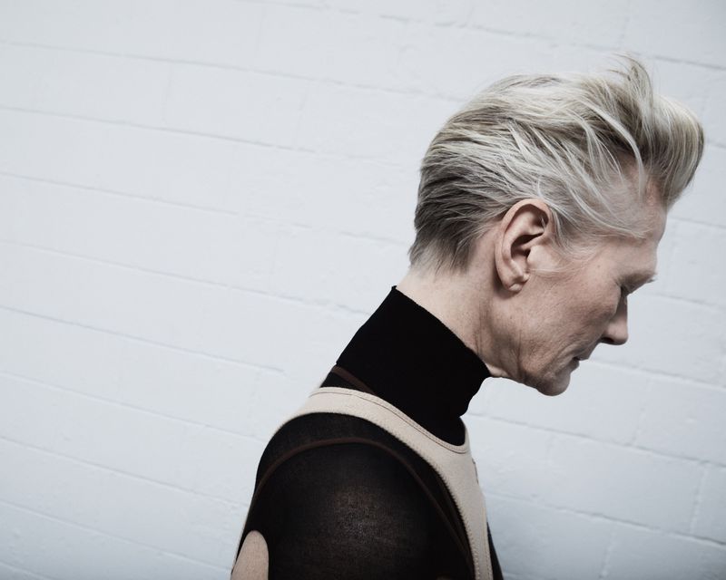 Profile portrait of an older adult male with grey-blonde hair, wearing a black turtleneck and patterned sweater, against a white wall.