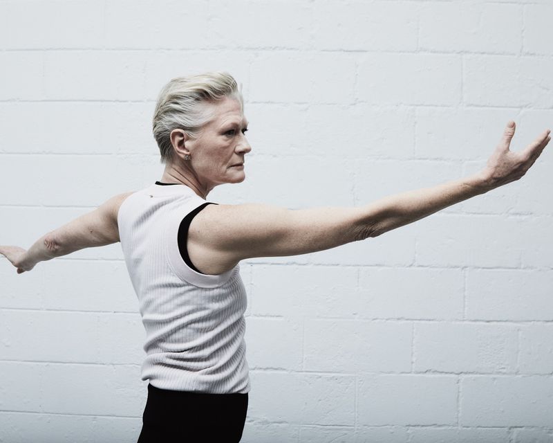 Older woman with short white hair and sleeveless shirt stands in front of white brick wall with arms outstretched in a dancer-like pose.
